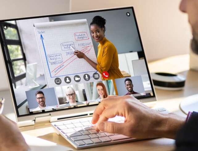 Foto. Eine Person sitzt vor einem Bildschirm auf dem eine Videokonferenz zu sehen ist. Die vortragende Person steht vor einem Flippboard und ist im Vollbildmodus zu sehen. Am unteren Rand sind vier weitere Kacheln mit Personen, die an der Konferenz teilnehmen.