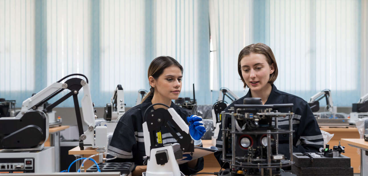Foto von 2 Personen in einer Werkstatt mit vielen verschiedenen Geräten. Vor ihnen steht ein Roboter