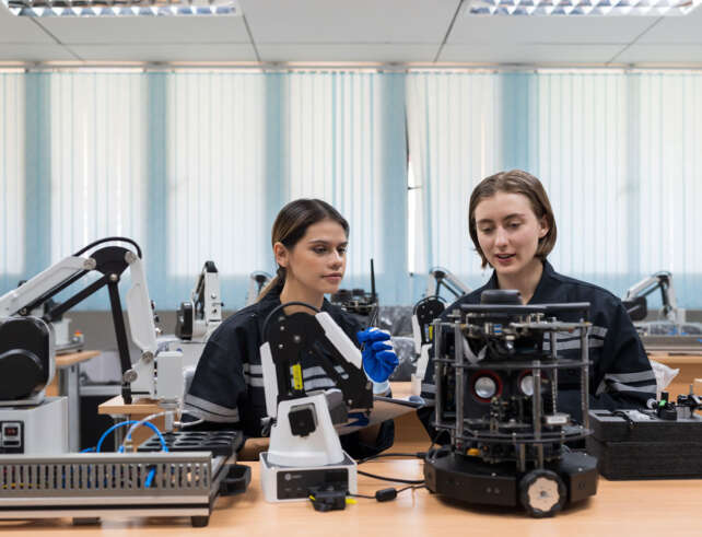 Foto von 2 Personen in einer Werkstatt mit vielen verschiedenen Geräten. Vor ihnen steht ein Roboter